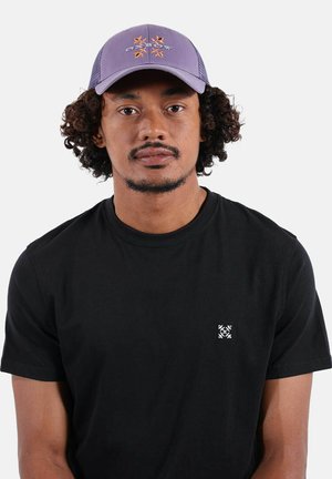 Homme aux cheveux bouclés portant une casquette violette avec un motif orange et un t-shirt noir avec un petit logo blanc sur la poitrine, faisant face à l'avant sur un fond blanc.