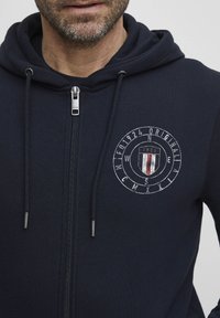 Sweat à capuche bleu marine zippé en tissu doux, avec un design graphique circulaire comprenant un blason et des accents rouge, blanc et bleu.