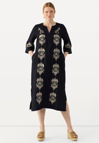 Das navyblaue Kleid zeichnet sich durch aufwendige goldene Stickereien mit floralen Mustern, Dreiviertelärmeln und Seitentaschen aus und wird mit braunen Plateau-Schuhen kombiniert.