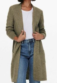 Olivově zelený pletený kardigan s dlouhým siluetu, otevřeným předním dílem, dvěma předními kapsami a vroubkovanými manžetami, nošený přes bílé tričko a modré džíny.