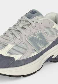 Γκρι και λευκό αθλητικό παπούτσι New Balance με πλέγμα, σουέτ επικαλύψεις, γκρι κορδόνια και χοντρή επενδεδυμένη σόλα, γωνιακά για να εμφανίζει το πλάι και την κορυφή.