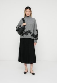 Pull gris avec des broderies florales noires à l'ourlet, associé à une jupe plissée noire et des chaussures plates. Le mannequin tient un sac noir.