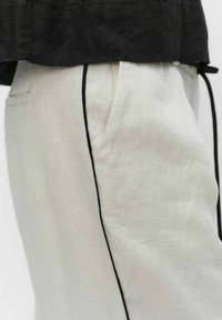 Lichtbeige linnen broek met zwarte bies langs de zijnaad, gedragen met een zwart topje dat deels boven de taille zichtbaar is.