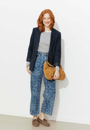 Femme rousse souriante portant une chemise à rayures, un blazer bleu marine, un jean large à motif, des mocassins marron, tenant un sac à main nude clouté contre un mur blanc.