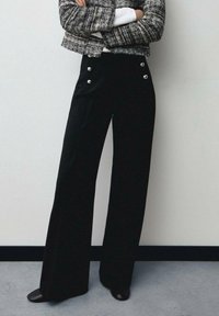 Un pantalon large noir en tissu lisse, avec de grands accents de boutons argentés et des poches avant, associé à une veste grise texturée.