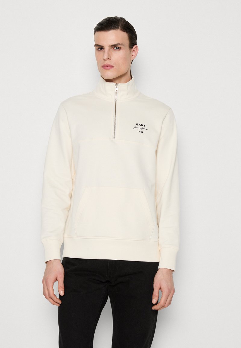 Pullover crema a zip con tasca frontale, caratterizzato da un logo ricamato a contrasto e polsini a coste. Tessuto morbido e testurizzato.