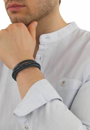 Mężczyzna w białej koszuli ze stójką, opierający podbródek na dłoni, noszący na lewym nadgarstku czarną plecioną skórzaną bransoletę.