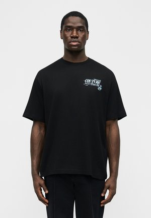 Muž s neutrlným výrazom tváre, oblečený v čiernom nadrozmernom tričku s malým bielym nápisom "Couture Beach Club" na ľavej strane hrude, stojaci pred jednofarebným pozadím.