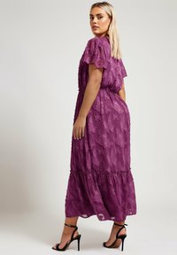 Lång lila klänning i strukturerad tyg med blommönster, korta svävande ärmar, elastisk midja och en tiered fåll. Svarta högklackade sandaler.