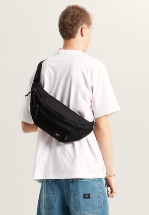 Zwarte schoudertas gemaakt van duurzaam stof, met ritsen, mesh accenten en een minimalistisch ontwerp, over het lichaam gedragen boven een wit T-shirt.