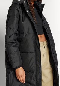 Černá prošívaná bunda s kapucí, zapínáním na zip, knofl�íkovými manžetami a prošívanými částmi. Hladká látka s matným povrchem.