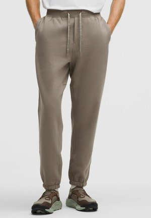 Mand iført taupe joggers med snoretræk og brune sneakers, hænderne i lommerne, stående mod en ensfarvet lys baggrund.