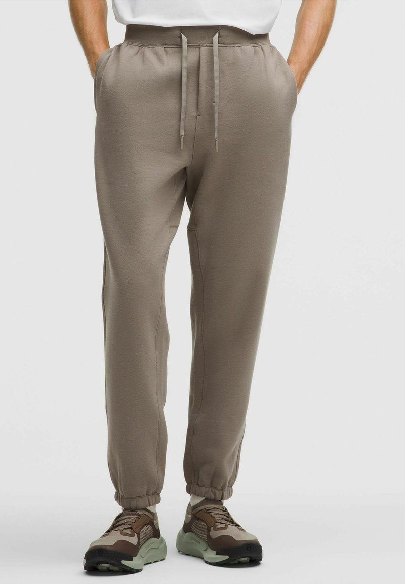 Man i taupe drawstring joggers och bruna sneakers, med händerna i fickorna, stående mot en ljus, enfärgad bakgrund.