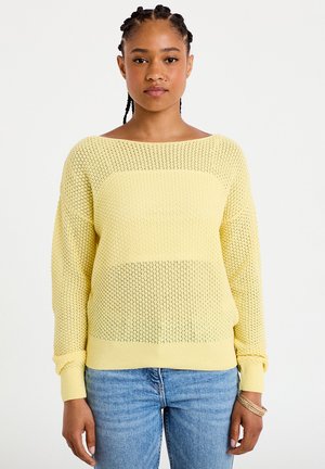 Vrouw met gevlochten haar die een gele gebreide trui en blauwe spijkerbroek draagt, staand tegen een effen witte achtergrond.