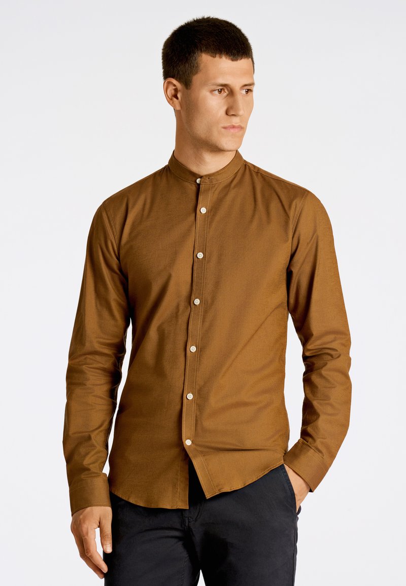 Camisa marrón de manga larga con cuello alzado, cierre de botones en la parte delantera y un ajuste entallado. Tejido suave con una textura sutil, llevada sobre pantalones oscuros.