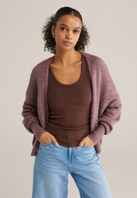 Cardigan tricotat mov pal, cu față deschisă, manșete cu dungi și croială lejeră, asortat cu un maiou maro și blugi evazați de culoare albastru deschis.