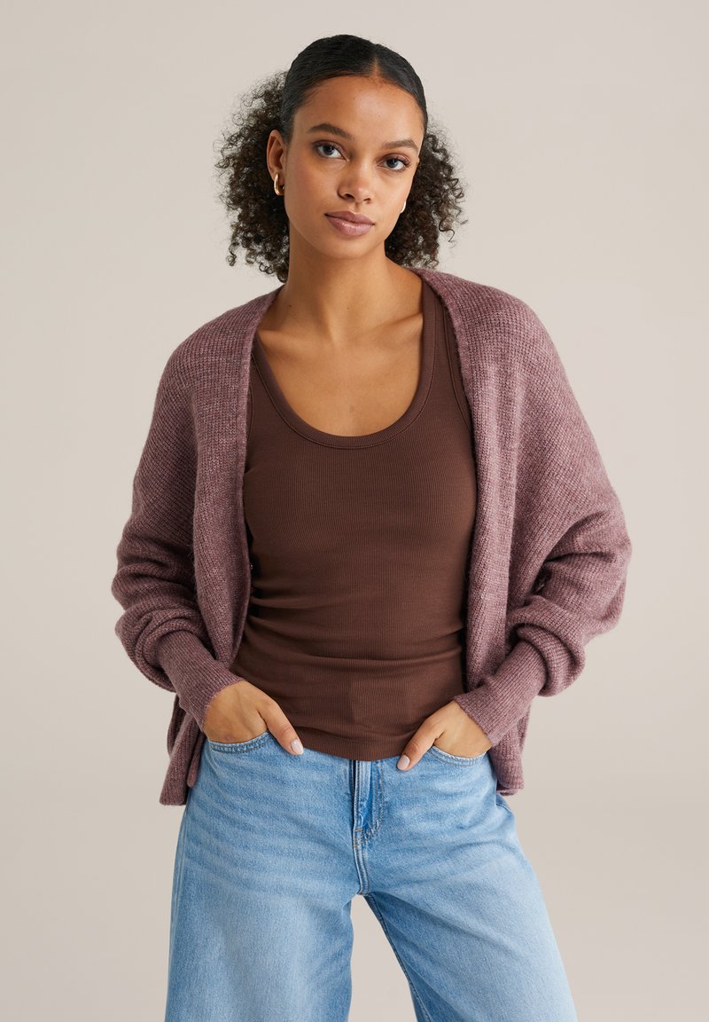 Cardigan tricotat mov pal, cu față deschisă, manșete cu dungi și croială lejeră, asortat cu un maiou maro și blugi evazați de culoare albastru deschis.