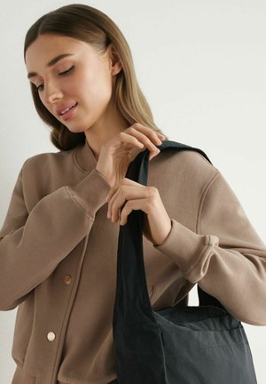 Kobieta w beżowej kurtce trzyma i dopasowuje pasek dużej czarnej torby na ramię na tle jednolitego jasnego tła.