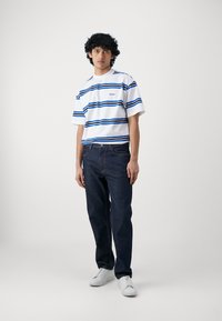 T-shirt bianca a righe blu, abbinata a jeans in denim scuro e sneakers bianche. Vestibilità casual con un design moderno.