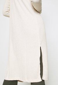 Vestido beige acanalado que llega hasta la mitad del muslo con aberturas laterales, tejido texturizado, dobladillo redondeado y mangas ajustadas, combinado con leggings verdes oliva.
