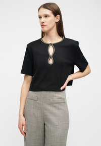 Mujer con camiseta negra de manga corta y detalle de escote en forma de ojo de cerradura, y pantalones grises de talle alto y pierna ancha, de pie con la mano en la cadera.