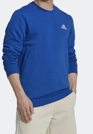 Blauwe sweatshirt met lange mouwen, ronde hals, zachte textuur en wit Adidas-logo op de linkerborst. Gedragen over beige broek.
