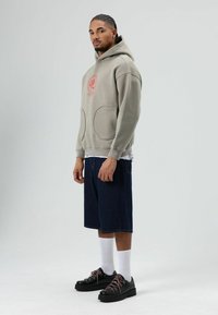 Sweat à capuche gris clair avec un motif circulaire rouge, grandes poches avant, porté avec un short en denim bleu marine et des chaussures noires aux lacets roses.