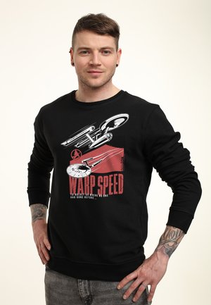 Mężczyzna ubrany w czarną bluzę z grafiką statku kosmicznego i czerwonym napisem „Warp Speed” oraz szare dżinsy, stoi z rękami na biodrach.