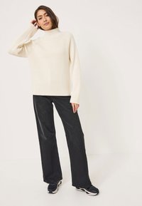 Cream ribbed turtleneck sweater with long sleeves paired with loose-fitting dark gray pants. Model wears black sneakers. Simple design.