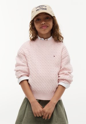 Ung person iført en lyserød kabelstrikket sweater, grøn nederdel, hvide opkørte skjorteærmer og en beige Tommy Hilfiger kasket.