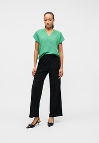 Γυναίκα που στέκεται φορώντας πράσινο τοπ με κοντά μανίκια και V-λαιμόκοψη, μαύρο παντελόνι με φαρδιά πόδια και μαύρα μυτερά παπούτσια με λουράκι στη φτέρνα.