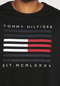 Černé tričko s vodorovnými pruhy v červené, bílé a námořnické barvě, s nápisem "TOMMY HILFIGER" a "EST. MCMLXXXV" nad pruhy.