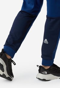 Granatowe spodnie sportowe z teksturowanym panelem na kolanach i logo, w połączeniu z czarno-białymi trampkami z bieżnikiem i akcentami marki.