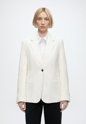 Donna con capelli biondi corti che indossa un blazer bianco sopra una camicia bianca con colletto e pantaloni neri, rivolta in avanti su uno sfondo semplice.