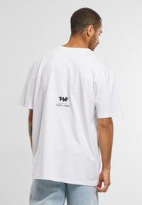 Hvit bomulls t-skjorte med løs passform, med en svart logo på ryggen. Korte ermer og rund hals, vist fra baksiden.