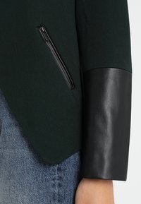 Zelený látkový blazer s černým koženým manžetou; má zipovou kapsu na levé straně, čisté linie a dobře padnoucí siluetu.