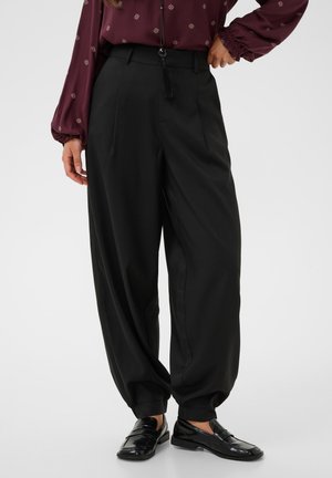 Femme portant un pantalon noir taille haute à jambes larges avec des revers aux chevilles, un chemisier bordeaux à motif et des mocassins noirs brillants.