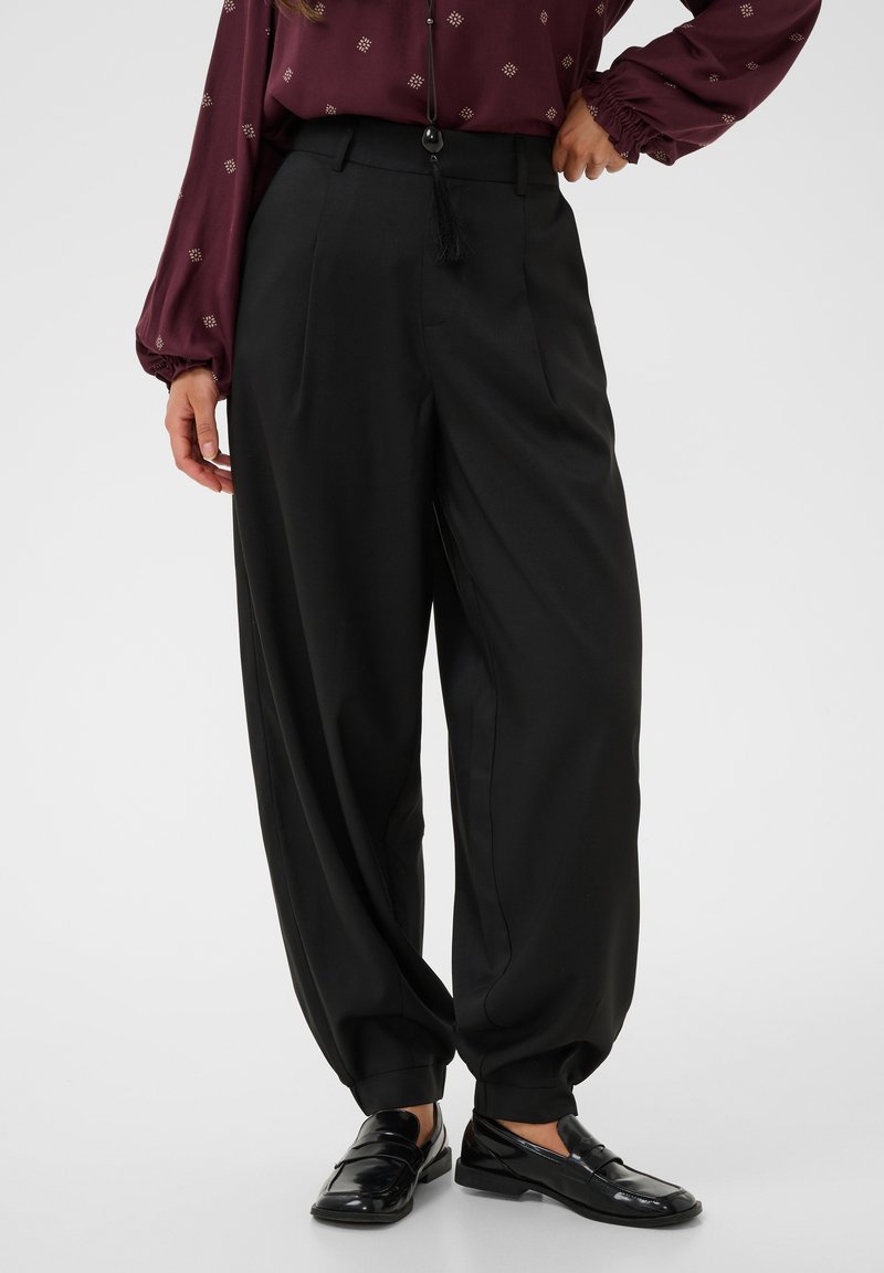 Femme portant un pantalon noir taille haute à jambes larges avec des revers aux chevilles, un chemisier bordeaux à motif et des mocassins noirs brillants.