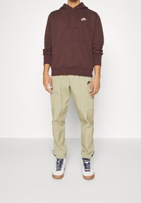 Brun luvtröja med vitt Nike-logotyp, matchad med ljus khaki cargobyxor med fickor och svart Nike-logotyp, samt vita sneakers.