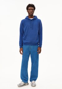 Blauer Kapuzenpullover aus Baumwolle mit einer Fronttasche und Kordelzug an der Kapuze, kombiniert mit hellblauen Jeans und grauen Sneakers mit strukturiertem Design.