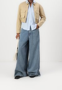 Henkilö, jolla on päällään vaaleanbeige lyhyt takki, vaaleansininen raidallinen paita, leveät farkut, mustat kengät ja valkoinen käsilaukku.