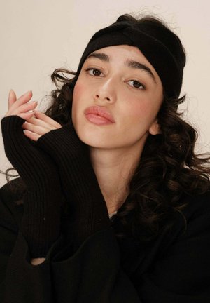 Une jeune femme aux cheveux bouclés porte un serre-tête noir et des gants sans doigts noirs, reposant ses mains près de son visage avec une expression neutre.