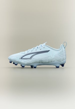 Λευκό ποδοσφαιρικό παπούτσι με ναυτικό μπλε καρφιά στη σόλα και ένα ναυτικό μπλε, επιμηκυμένο τριγωνικό σχέδιο στο πλάι, πάνω σε απλό μπεζ φόντο.