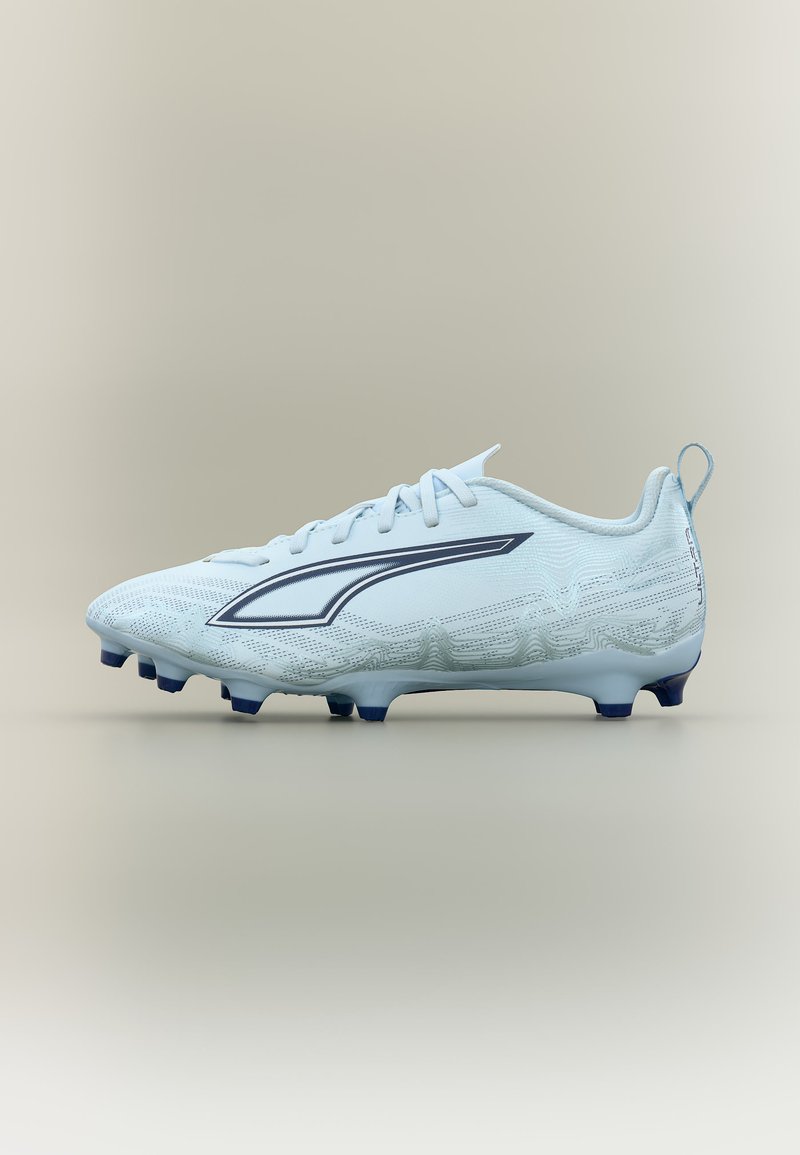 Chaussure de soccer blanche avec des crampons bleu marine et un design triangulaire allongé bleu marine sur le côté, sur un fond beige uni.