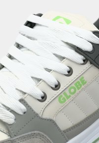 Gray and cream sneaker with white laces. Features a green logo and gray accents. Made of smooth synthetic materials with textured panels.