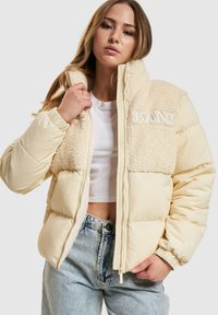 Beige puffer-jacka med en fluffig, krämfärgad sherpa-panel. Har en dragkedja framtill och detaljer med broderad "KANI". Bärs över en vit topp.