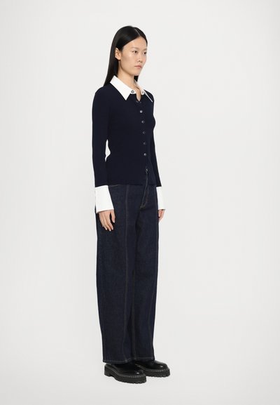 Marineblauwe geribbelde cardigan met een witte kraag, knopen aan de voorkant en contrasterende mouwen, gecombineerd met losse donkere denim jeans en zwarte platform schoenen.