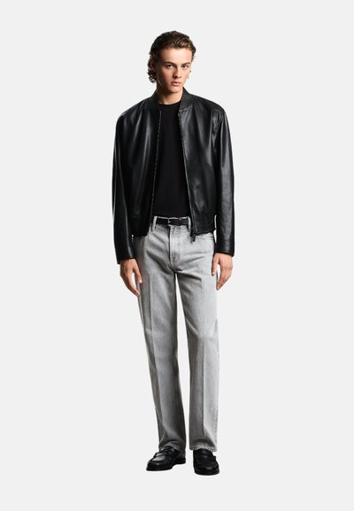 Giacca bomber in pelle nera sopra una t-shirt nera a coste, abbinata a jeans grigi chiari e scarpe nere. Il modello è posizionato su uno sfondo bianco.