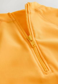 Dzeltens zip-up apģērbs no gluda materiāla ar īsu rāvējslēdzēju. Aprīkots ar plakaniem šuvēm un augstu apkakli.