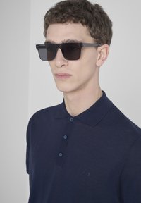 Occhiali da sole rettangolari neri indossati da una persona, con una polo blu navy caratterizzata da tre bottoni e un logo discreto sul petto.